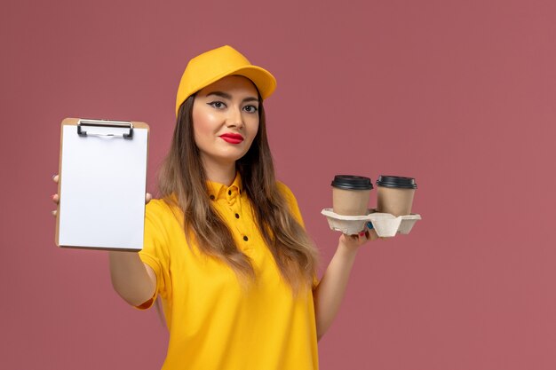 Vue de face du courrier féminin en uniforme jaune et cap tenant le bloc-notes de tasses de café de livraison souriant sur mur rose clair
