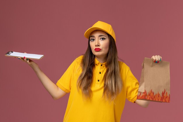 Vue de face du courrier féminin en uniforme jaune et cap tenant le paquet de nourriture et le bloc-notes sur le mur rose