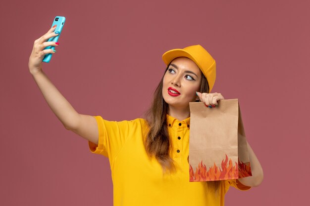 Vue de face du courrier féminin en uniforme jaune et cap tenant le paquet de nourriture et prenant un selfie sur le mur rose