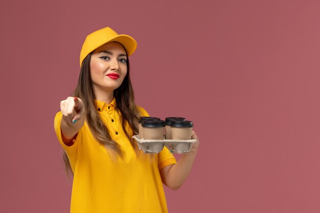 Vue de face du courrier féminin en uniforme jaune et cap tenant des tasses de café de livraison marron sur le mur rose