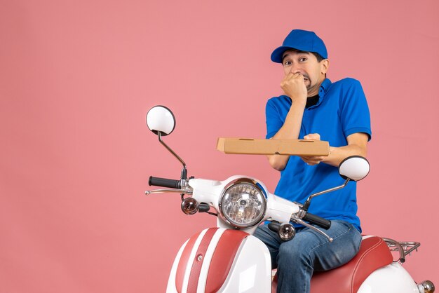 Vue de face du courrier homme portant un chapeau assis sur un scooter, peur sur fond de pêche pastel