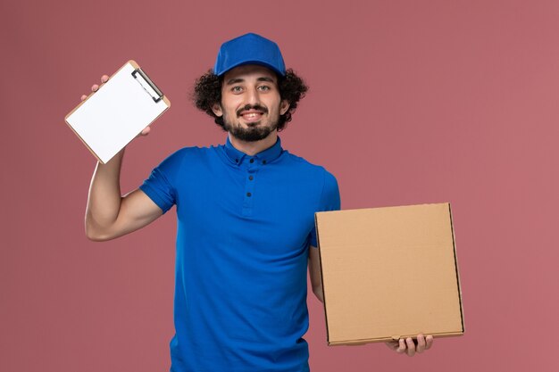 Vue de face du courrier masculin en chapeau uniforme bleu avec bloc-notes et boîte de nourriture sur ses mains sur le mur rose