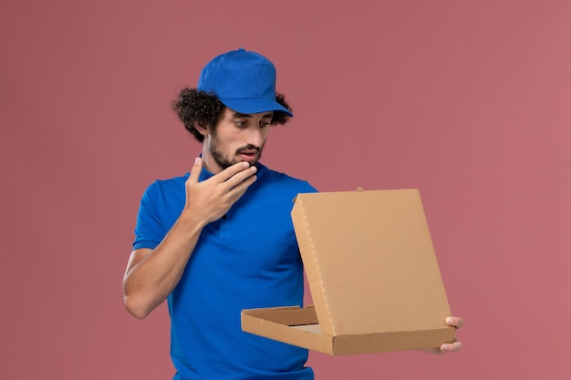 Vue de face du courrier masculin en chapeau uniforme bleu avec boîte de nourriture ouverte sur ses mains sur le mur rose clair