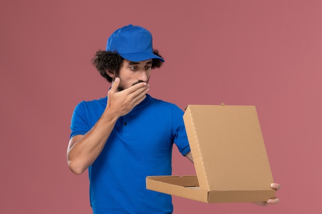 Vue de face du courrier masculin en chapeau uniforme bleu avec boîte de nourriture ouverte sur ses mains sur le mur rose clair