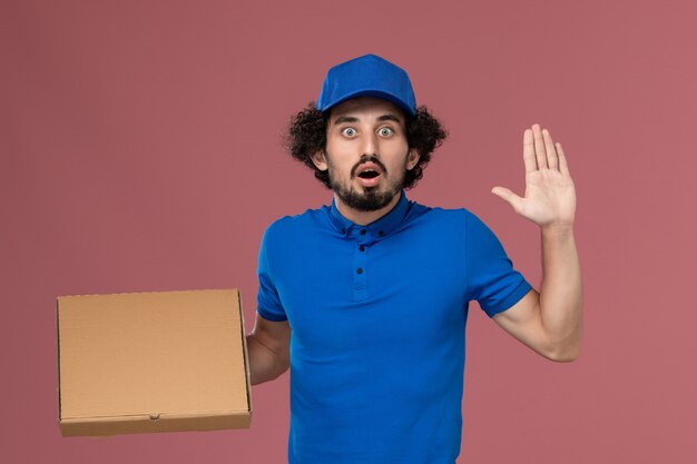 Vue de face du courrier masculin en chapeau uniforme bleu avec boîte de nourriture sur ses mains sur le mur rose clair