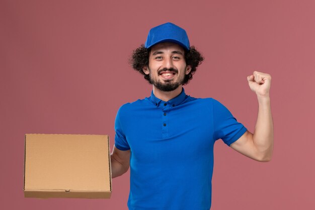 Vue de face du courrier masculin en chapeau uniforme bleu avec boîte de nourriture sur ses mains se réjouissant sur mur rose clair