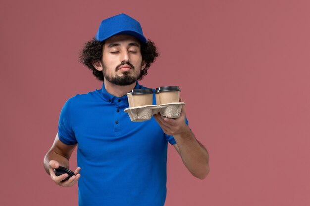 Vue de face du courrier masculin en chapeau uniforme bleu avec des tasses de café de livraison sur ses mains sur un mur rose clair