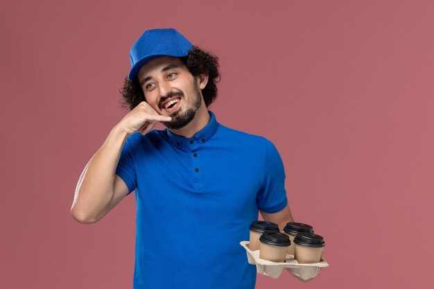 Vue de face du courrier masculin en chapeau uniforme bleu avec des tasses de café de livraison sur ses mains sur un mur rose clair