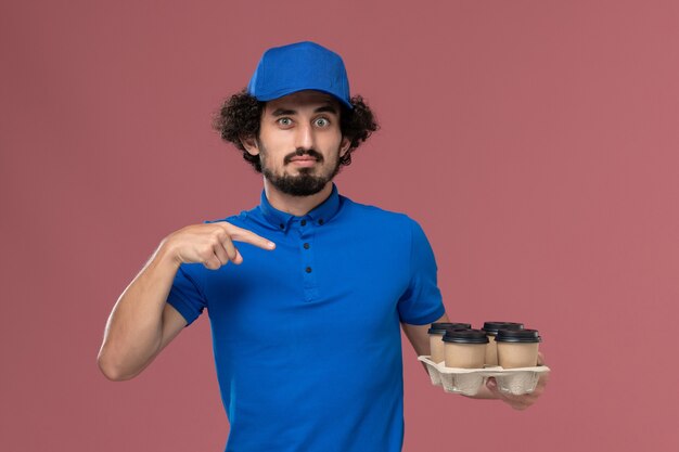 Vue de face du courrier masculin en chapeau uniforme bleu avec des tasses de café de livraison sur ses mains sur un mur rose clair