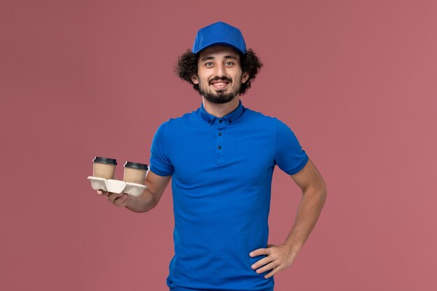Vue de face du courrier masculin en chapeau uniforme bleu avec des tasses de café de livraison sur ses mains sur le mur rose