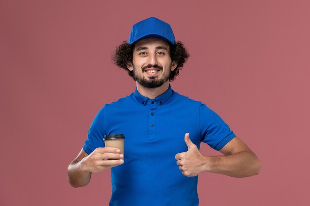 Vue de face du courrier masculin en uniforme bleu et capuchon avec tasse de café de livraison sur ses mains sur le mur rose