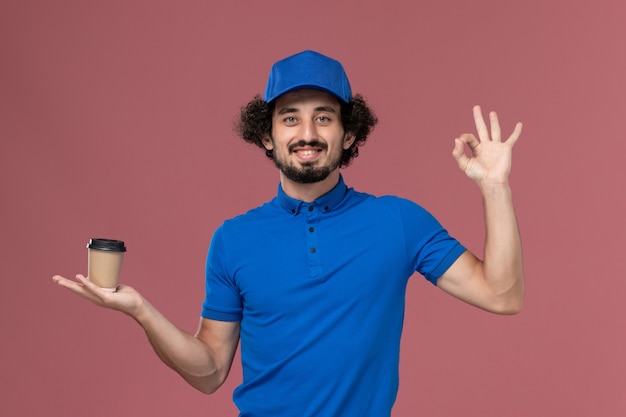 Vue de face du courrier masculin en uniforme bleu et capuchon avec tasse de café de livraison sur ses mains sur le mur rose