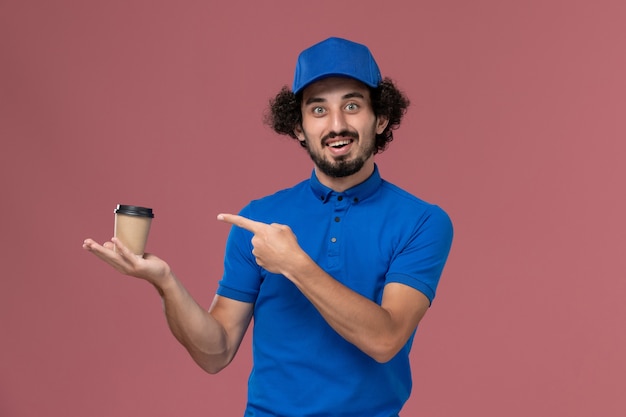 Vue de face du courrier masculin en uniforme bleu et capuchon avec tasse de café de livraison sur ses mains sur le mur rose