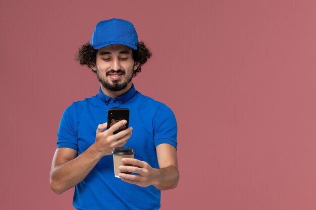 Vue de face du courrier masculin en uniforme bleu et capuchon avec tasse de café de livraison et smartphone sur ses mains sur le mur rose