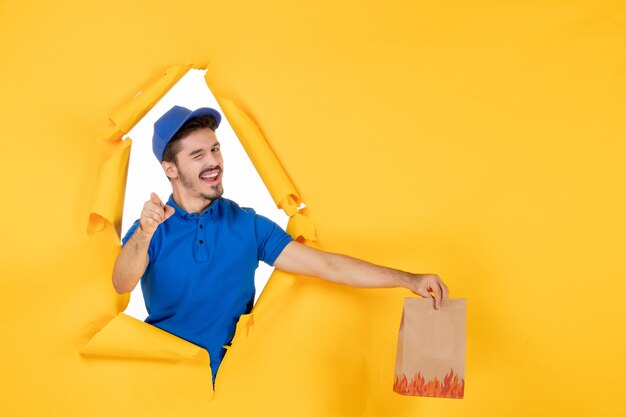 Photo gratuite vue de face du courrier masculin en uniforme bleu tenant un emballage de nourriture sur un espace jaune
