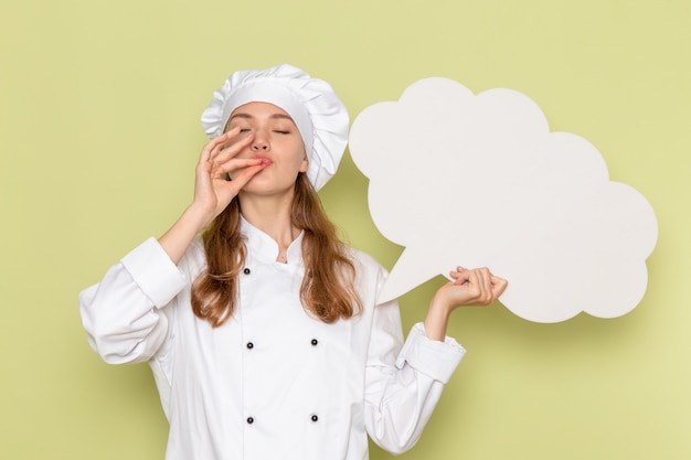 Vue de face du cuisinier en costume de cuisinier blanc tenant un énorme panneau blanc sur le mur vert