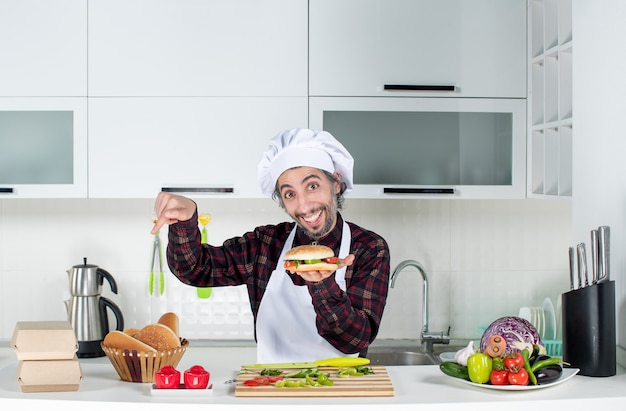 Vue de face du cuisinier masculin brandissant un hamburger pointant sur du pain sur une table debout derrière la table de la cuisine