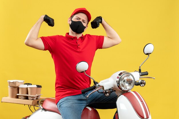 Vue de face du fier jeune adulte portant un chemisier rouge et des gants de chapeau en masque médical délivrant la commande assis sur un scooter montrant ses muscles sur fond jaune