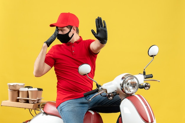 Vue de face du jeune adulte émotionnel portant un chemisier rouge et des gants de chapeau dans un masque médical délivrant la commande assis sur un scooter sur fond jaune