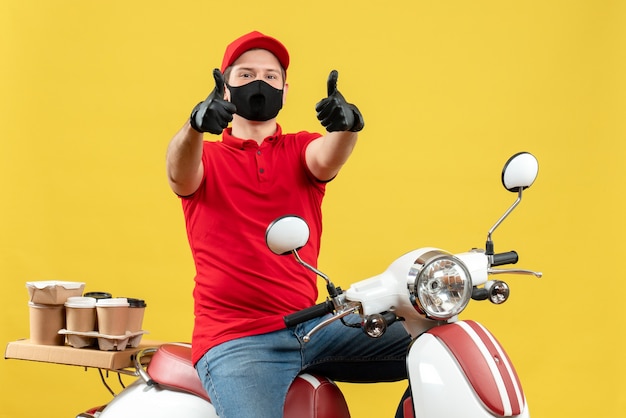 Photo gratuite vue de face du jeune adulte souriant portant un chemisier rouge et des gants de chapeau dans un masque médical délivrant la commande assis sur un scooter faisant un geste ok sur fond jaune