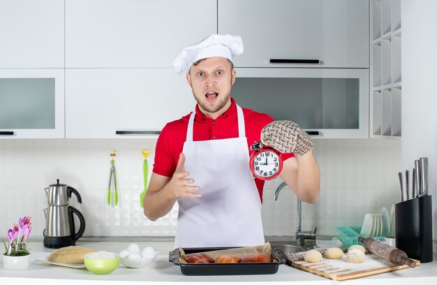 Vue de face du jeune chef masculin surpris portant un support tenant une horloge et faisant un geste correct dans la cuisine blanche