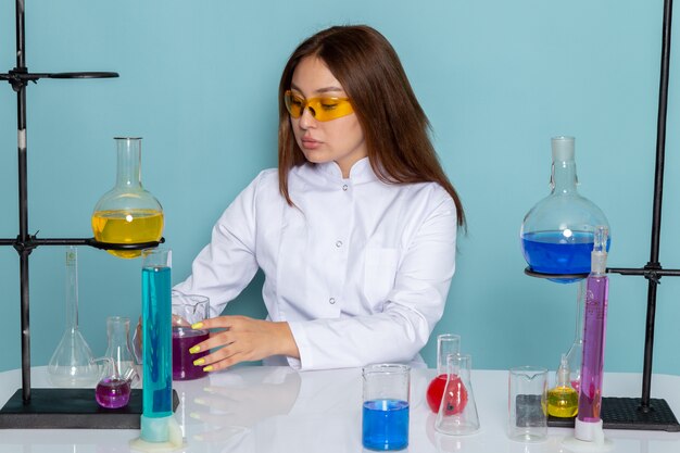 Vue de face du jeune chimiste féminin en costume blanc en face de table travaillant avec des solutions