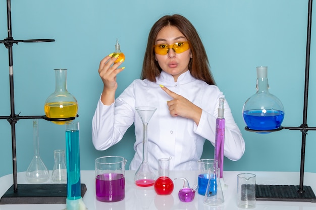 Vue de face du jeune chimiste féminin en costume blanc en face de table travaillant avec des solutions