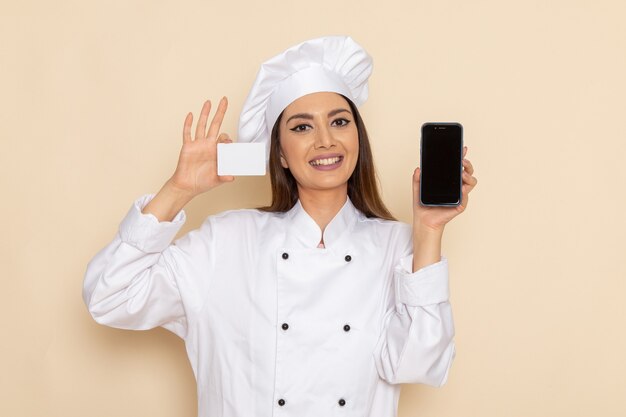 Vue de face du jeune cuisinier en costume de cuisinier blanc tenant la carte et le téléphone sur le mur blanc