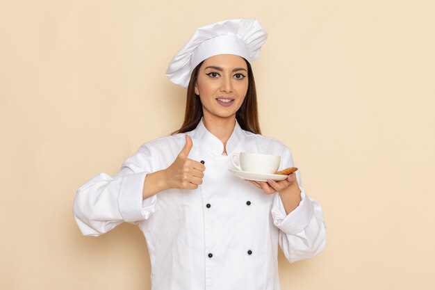 Vue de face du jeune cuisinier en costume de cuisinier blanc tenant une tasse de café et souriant sur un mur blanc
