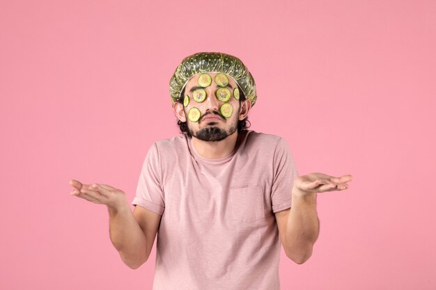 vue de face du jeune homme appliquant un masque de concombre sur son visage sur un mur rose