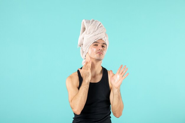 vue de face du jeune homme après la douche en appliquant de la crème sur son visage sur un mur bleu