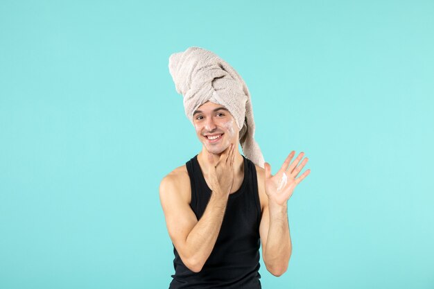 vue de face du jeune homme après la douche en appliquant de la crème sur son visage sur un mur bleu