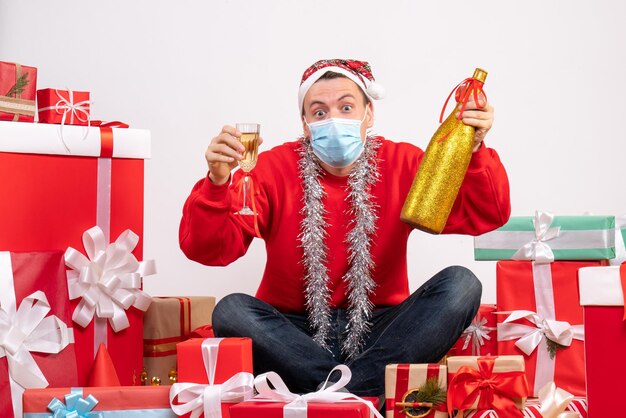 Vue de face du jeune homme assis autour de cadeaux célébrant avec du champagne sur un mur blanc