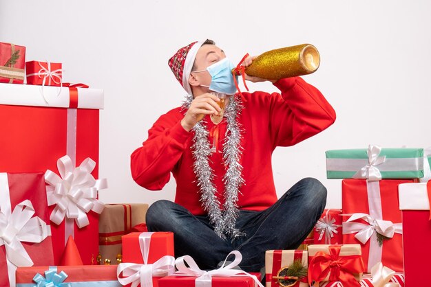 Vue de face du jeune homme assis autour de cadeaux célébrant avec du champagne sur un mur blanc