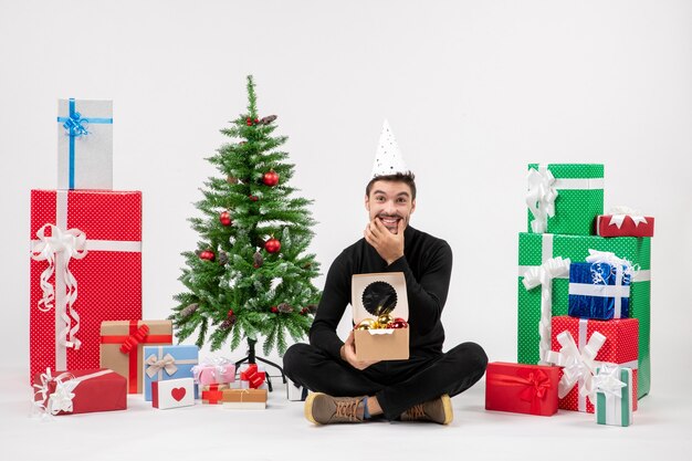 Vue de face du jeune homme assis autour de cadeaux sur un mur blanc