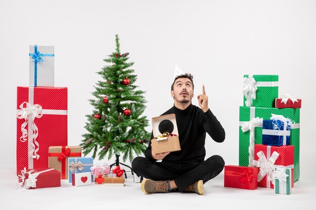 Vue de face du jeune homme assis autour de cadeaux sur un mur blanc