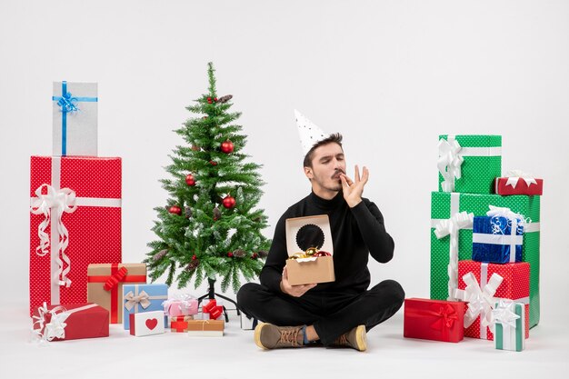 Vue de face du jeune homme assis autour de cadeaux sur un mur blanc