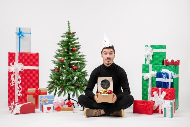 Vue de face du jeune homme assis autour de cadeaux sur un mur blanc