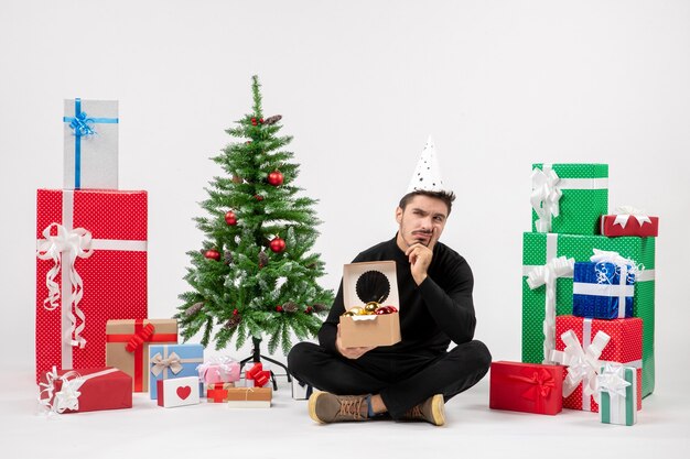 Vue de face du jeune homme assis autour de cadeaux sur un mur blanc