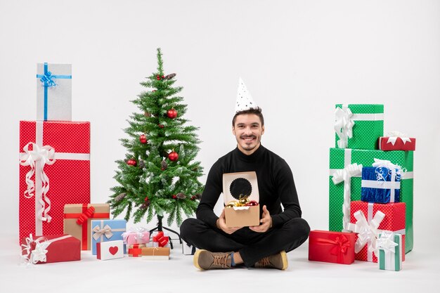 Vue de face du jeune homme assis autour de cadeaux sur un mur blanc