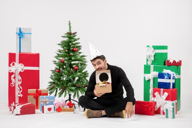 Vue de face du jeune homme assis autour de cadeaux sur un mur blanc