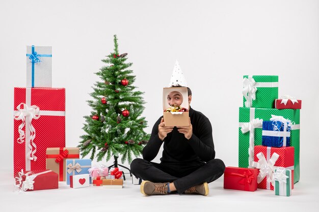 Vue de face du jeune homme assis autour de cadeaux sur un mur blanc