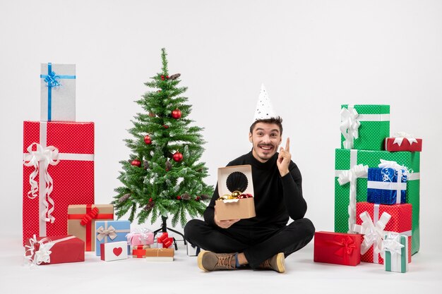 Vue de face du jeune homme assis autour de cadeaux sur un mur blanc