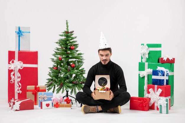 Vue de face du jeune homme assis autour de cadeaux sur un mur blanc