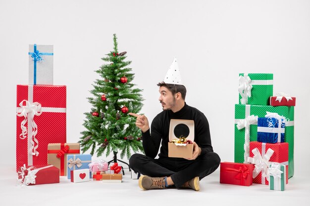 Vue de face du jeune homme assis autour de cadeaux sur un mur blanc