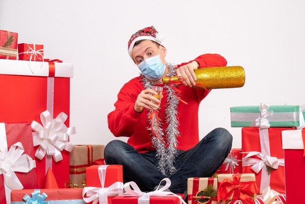 Vue de face du jeune homme assis autour de cadeaux de Noël célébrant avec du champagne sur un mur blanc