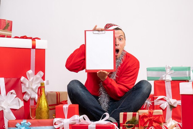 Vue de face du jeune homme assis autour de cadeaux de Noël sur mur blanc