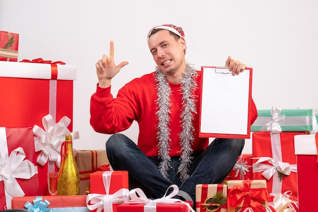 Vue de face du jeune homme assis autour de cadeaux de Noël avec presse-papiers sur mur blanc