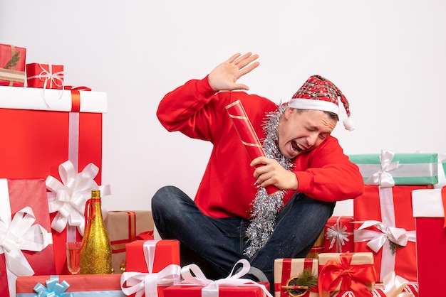 Vue de face du jeune homme assis autour de cadeaux avec pétard sur mur blanc