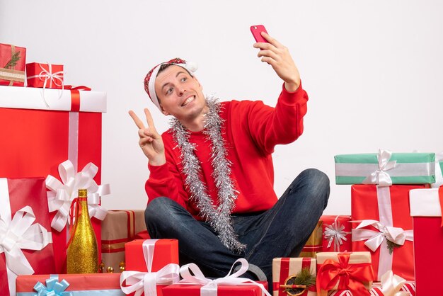 Vue de face du jeune homme assis autour de cadeaux prenant selfie sur mur blanc